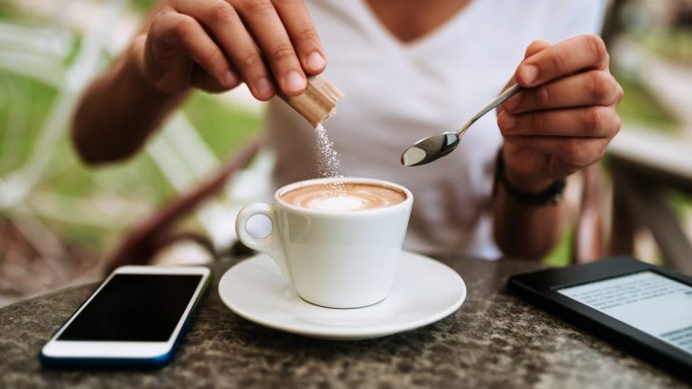 A person pouring a sugar packet into a latte in a white coffee cup.